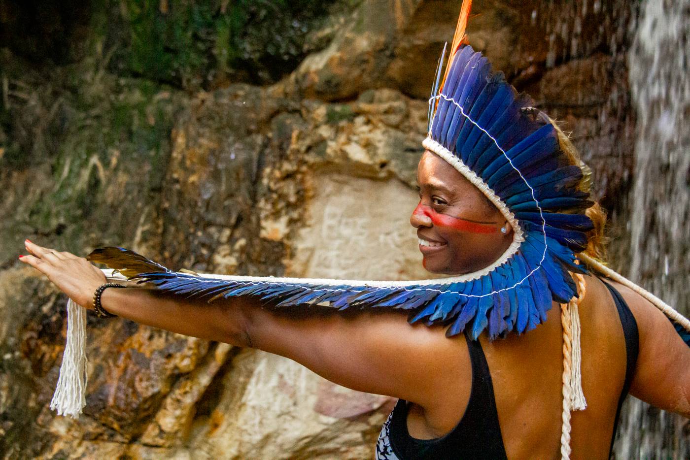 Mujer con penacho y alas de plumas azules extendiendo el brazo junto a la cascada