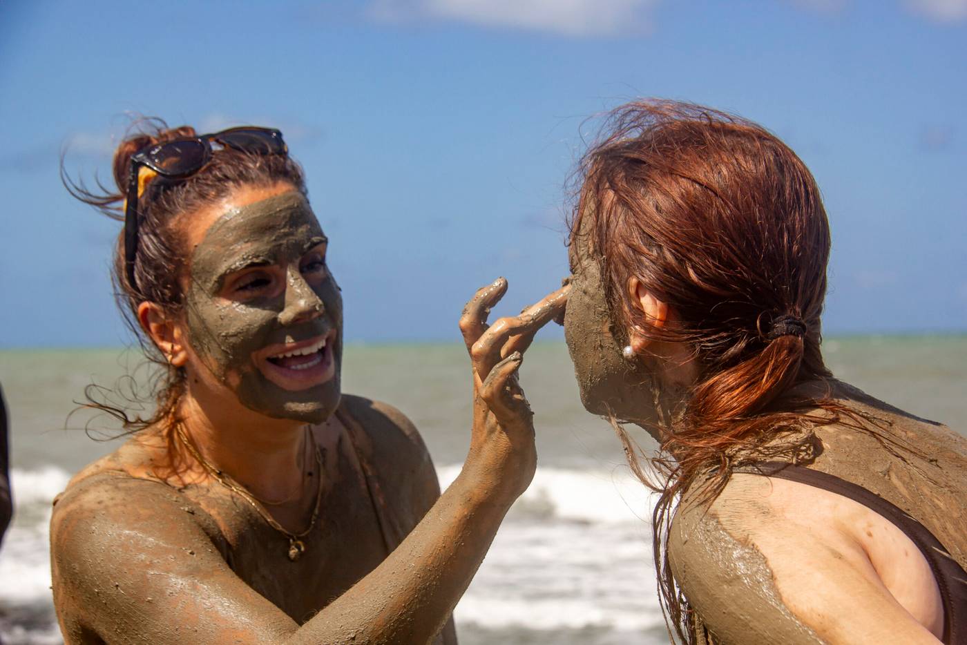 Dos mujeres riendo mientras se cubren la cara de arcilla junto al mar