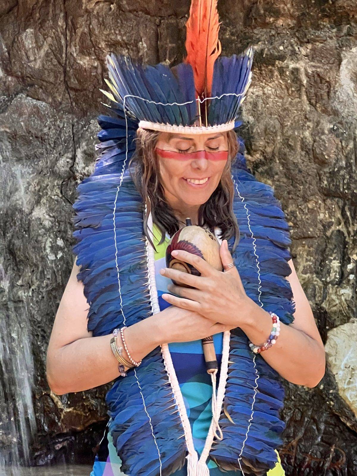 Mujer con penacho de plumas azules y los ojos cerrados sosteniendo una maraca sobre el pecho